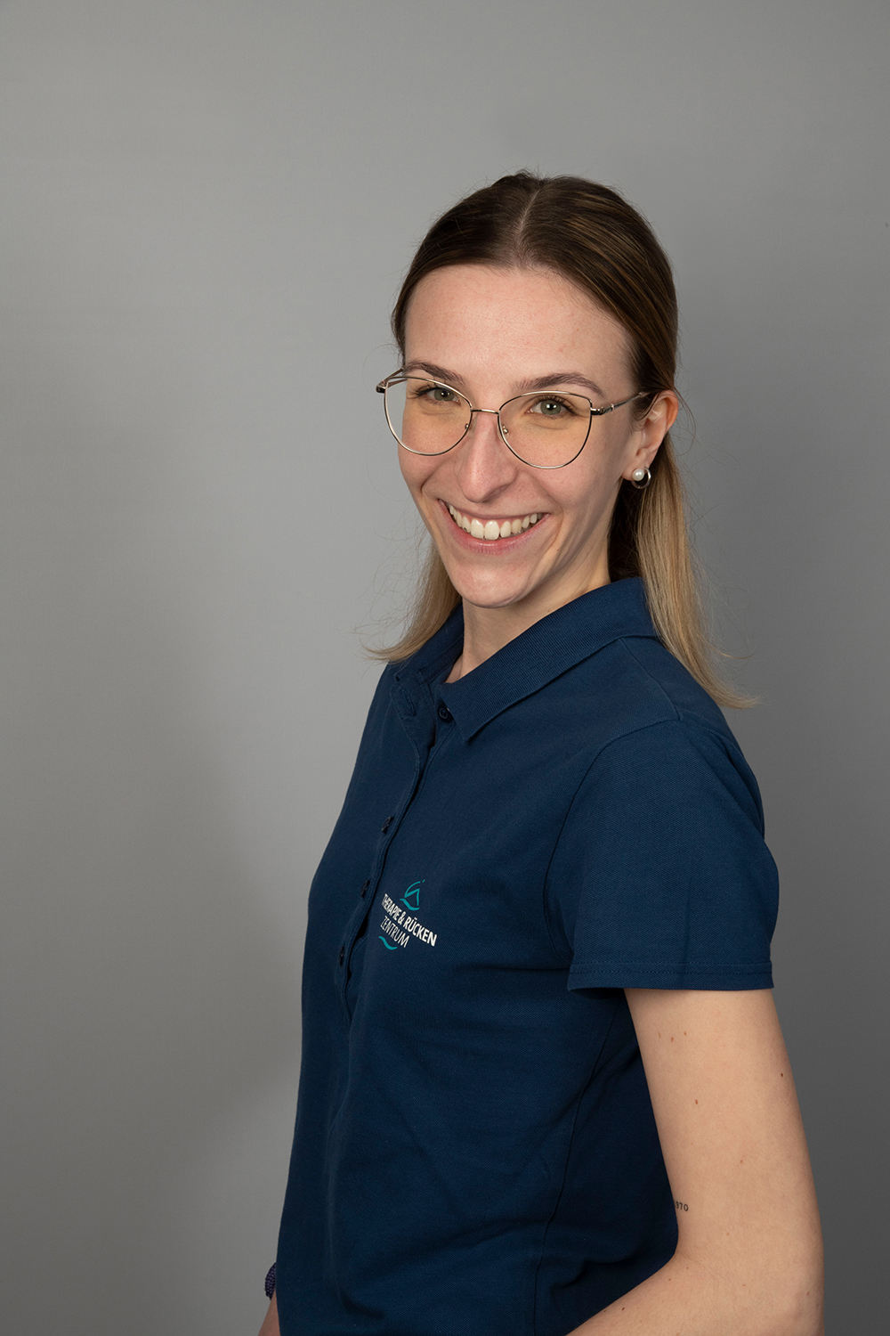 Eine Frau mit glattem, schulterlangem Haar und Brille lächelt in die Kamera. Sie trägt ein marineblaues Hemd mit Kragen und steht vor einem schlichten grauen Hintergrund.