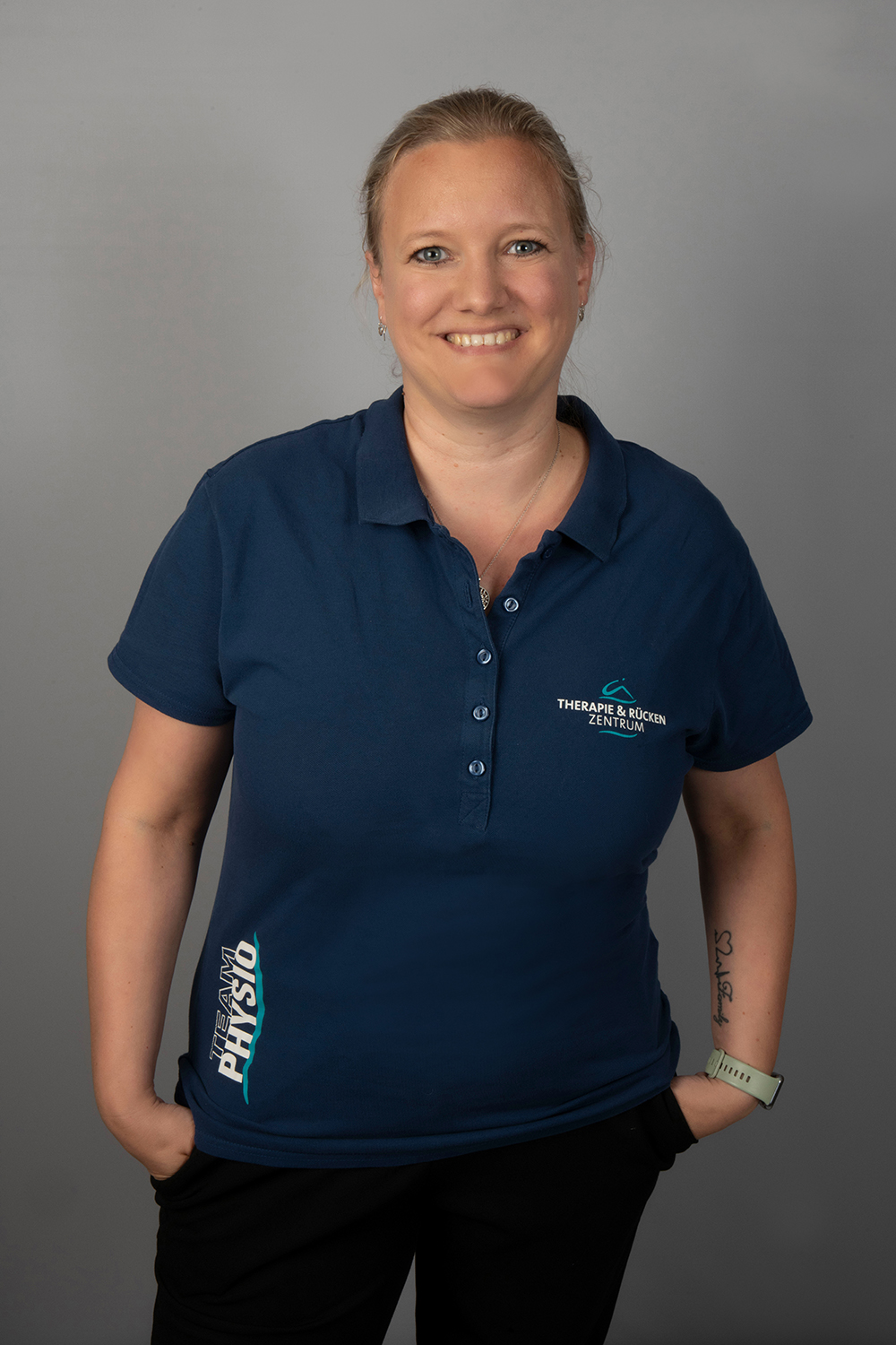 Eine lächelnde Frau steht vor einem grauen Hintergrund und trägt ein marineblaues Poloshirt mit dem Aufdruck "THERAPIE & RÜCKEN ZENTRUM" und "PHYSIO". Sie hat ihr helles Haar zurückgesteckt und trägt eine Smartwatch am Handgelenk.