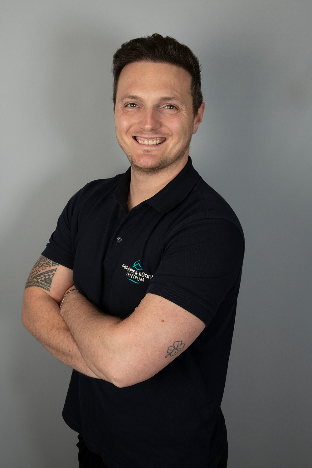 Ein lächelnder Mann mit kurzen dunklen Haaren steht mit verschränkten Armen da und trägt ein schwarzes Poloshirt mit einem Logo. Er hat sichtbare Tätowierungen auf seinen Armen und posiert vor einem schlichten hellgrauen Hintergrund.