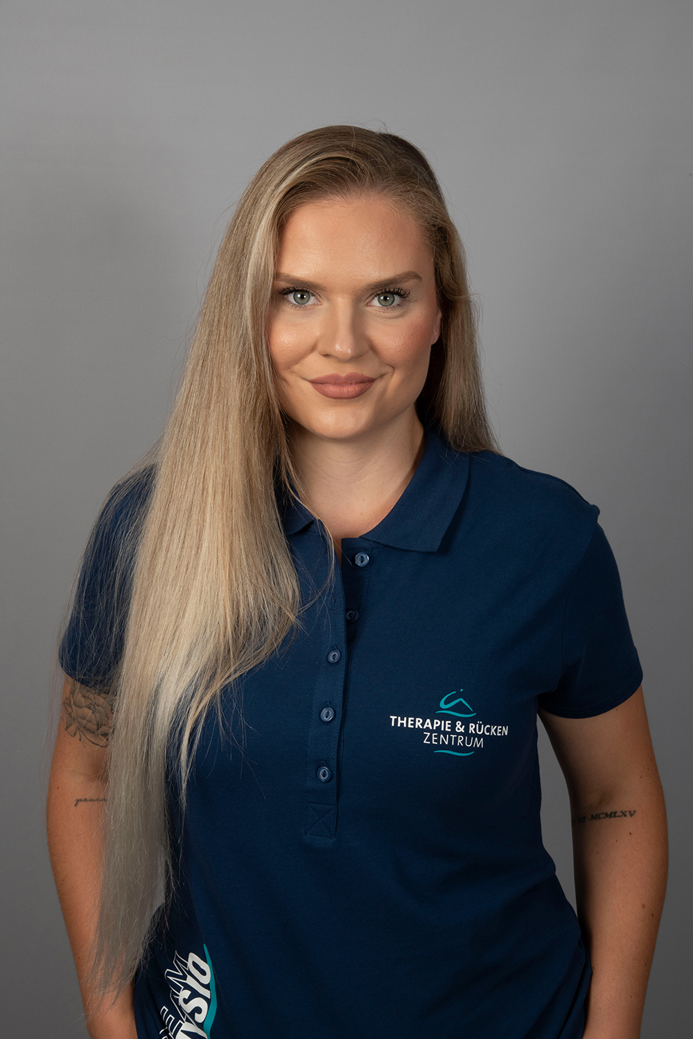 Eine Frau mit langen, glatten blonden Haaren und einer Tätowierung auf dem linken Arm trägt ein marineblaues Poloshirt mit dem Aufdruck THERAPIE & RÜCKEN ZENTRUM, steht vor einem einfarbig grauen Hintergrund und lächelt.