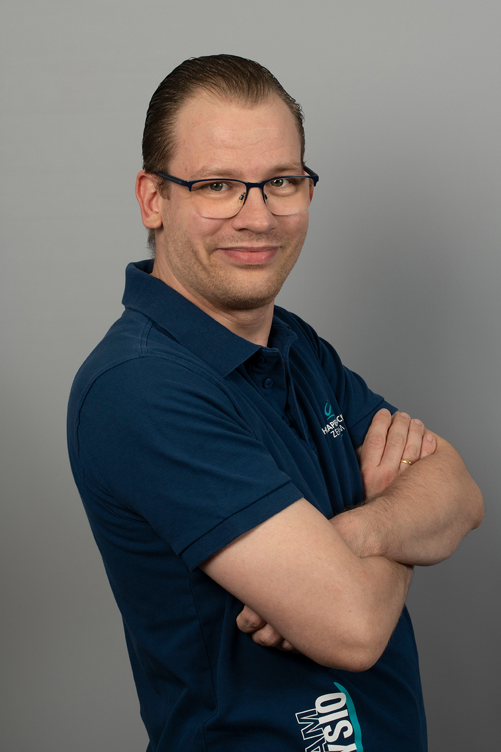 Ein Mann mit Brille und kurzem, zurückgekämmtem Haar steht vor einem grauen Hintergrund und lächelt mit verschränkten Armen. Er trägt ein dunkelblaues Poloshirt mit einem kleinen Logo auf der Brust.
